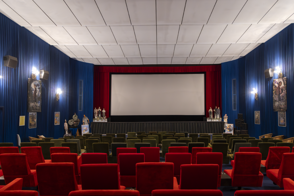 Der Saal des Kinos in Anspach. Rote und olivgrüne Sitze stehen in geraden Reihen, die Seitenwände sind mit blauem Stoff bespannt und mit Filmpostern und Aufstellern dekoriert. Vorn ist die Leinwand zu sehen.