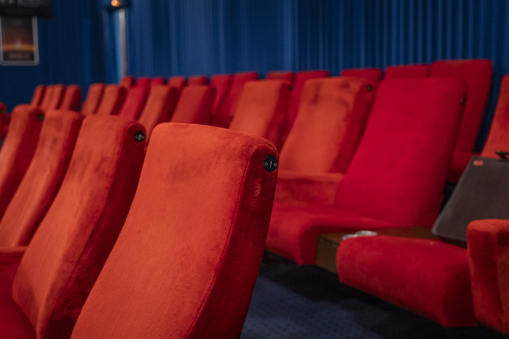 Nahaufnahme der Kinositze. Vorn ist die Lehne eines samtenen roten Sitzes zu sehen, an dem die Sitznummer 1 angebracht ist. Dahinter leicht unscharf die nächste Reihe roter Sitze.