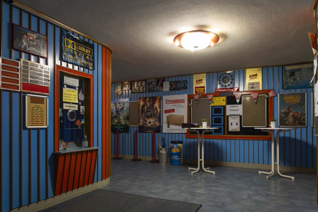 Foyer des Kinos Anspach. Die Wände sind mit rotem und blauem Holz getäfelt und mit Filmpostern behangen. Links ist der Ticket-Schalter zu sehen.