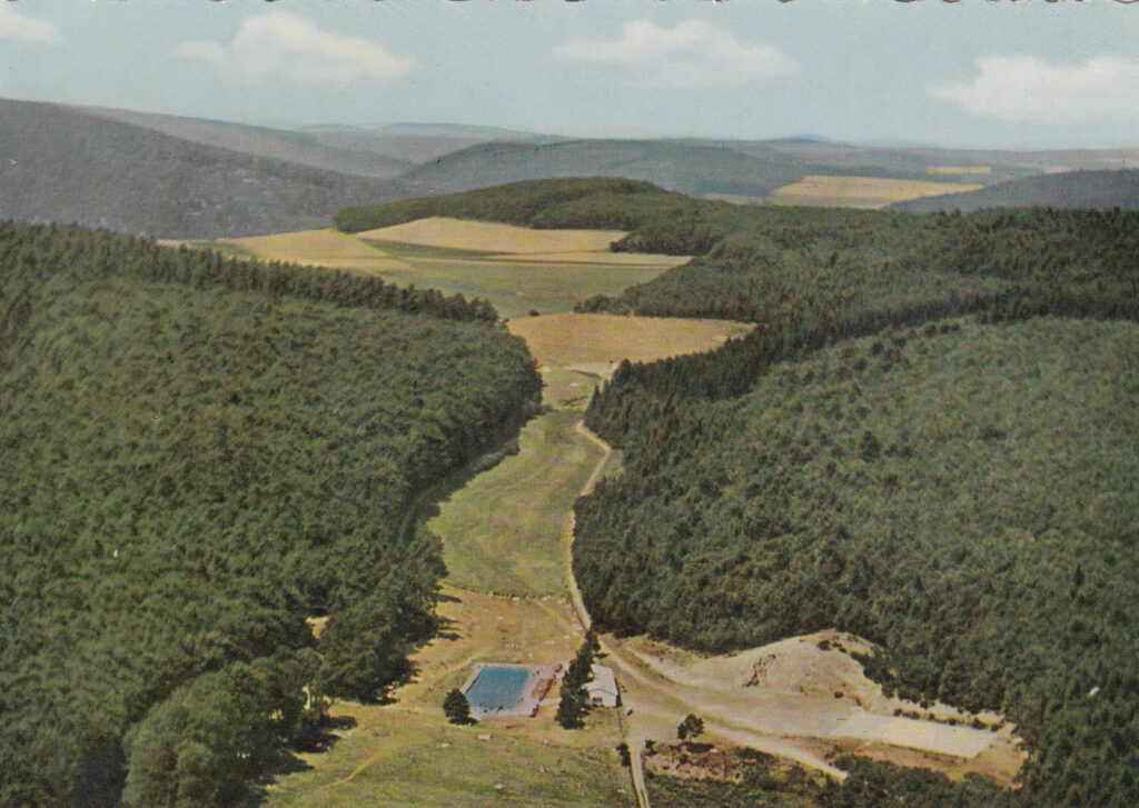 Alte Luftaufnahme des Waldschwimmbads. Zwischen bewaldeten Hügeln, Feldern und Wiesen ist das Becken zu sehen, das als kleines, türkisfarbenes Rechteck am unteren Bildrand liegt.