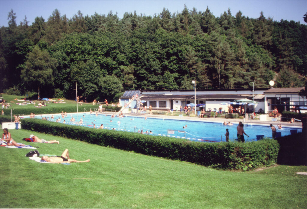 Ältere Aufnahme des Waldschwimmbads im Sommer. Im Wasser schwimmen Menschen, auf der Wiese liegen andere und sonnen sich.