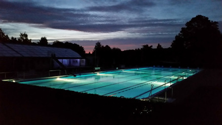 Aufnahme des Waldschwimmbads bei Nacht. Das Becken leuchtet in hellem Türkis, der Himmel ist vom Abendrot gefärbt.